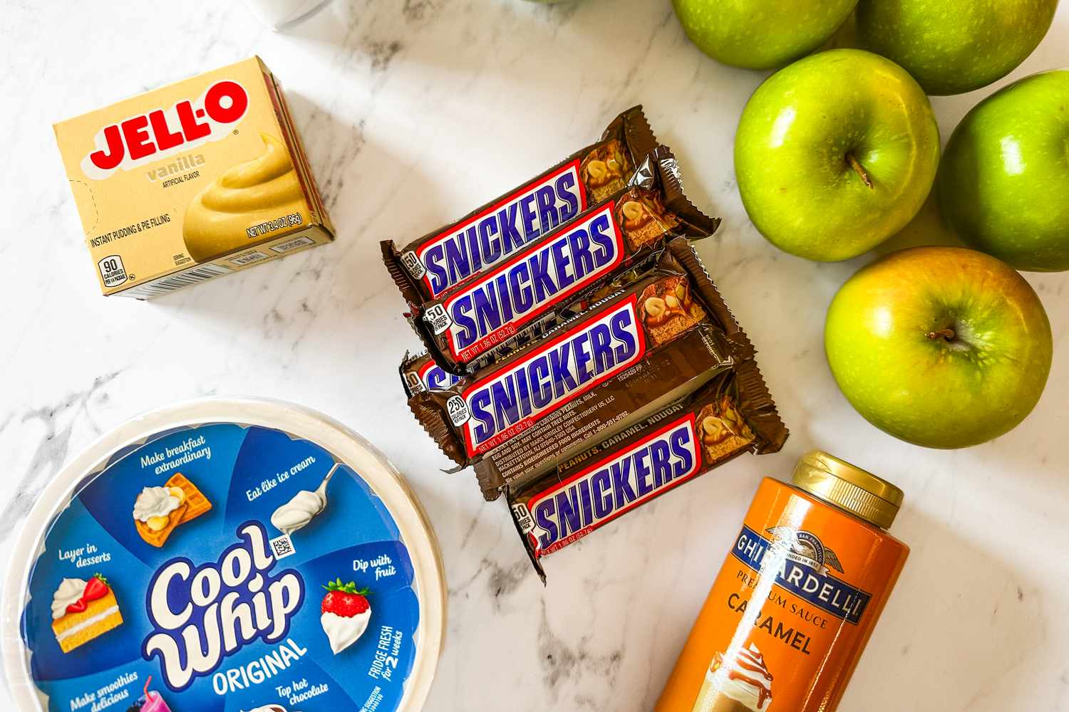 Ingredients for Snickers salad including apples, Snickers bars, Cool Whip, caramel sauce, and banana pudding mix