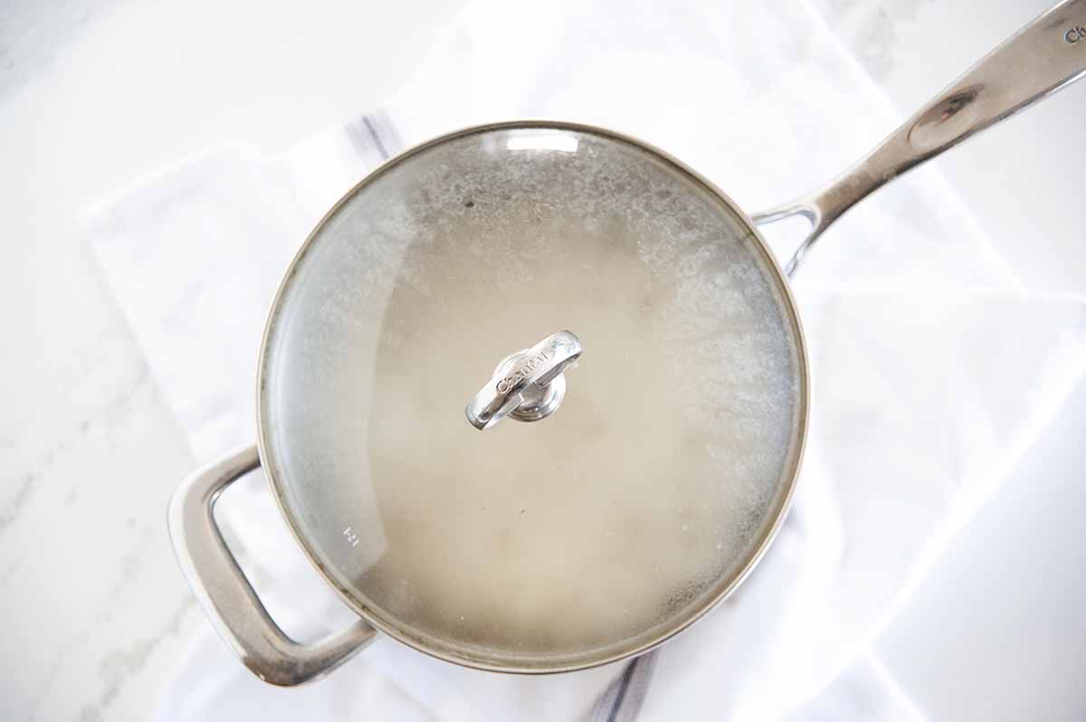 Saucepan with lid on and steamy white rice inside. The pot is resting on a white linen on a marble table.