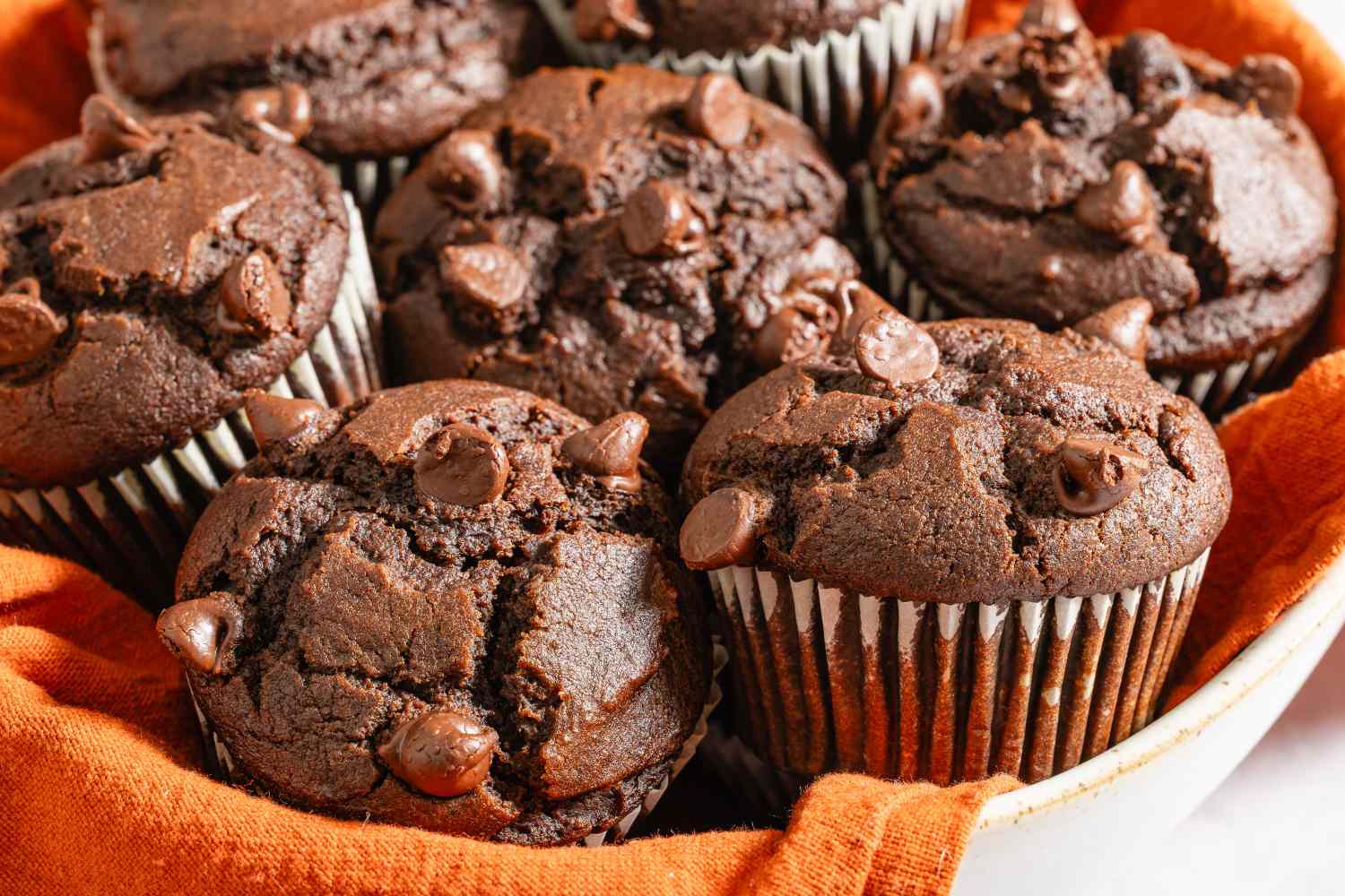 A plate of chocolate muffins with chocolate chips on top arranged in a bowl with an orange cloth