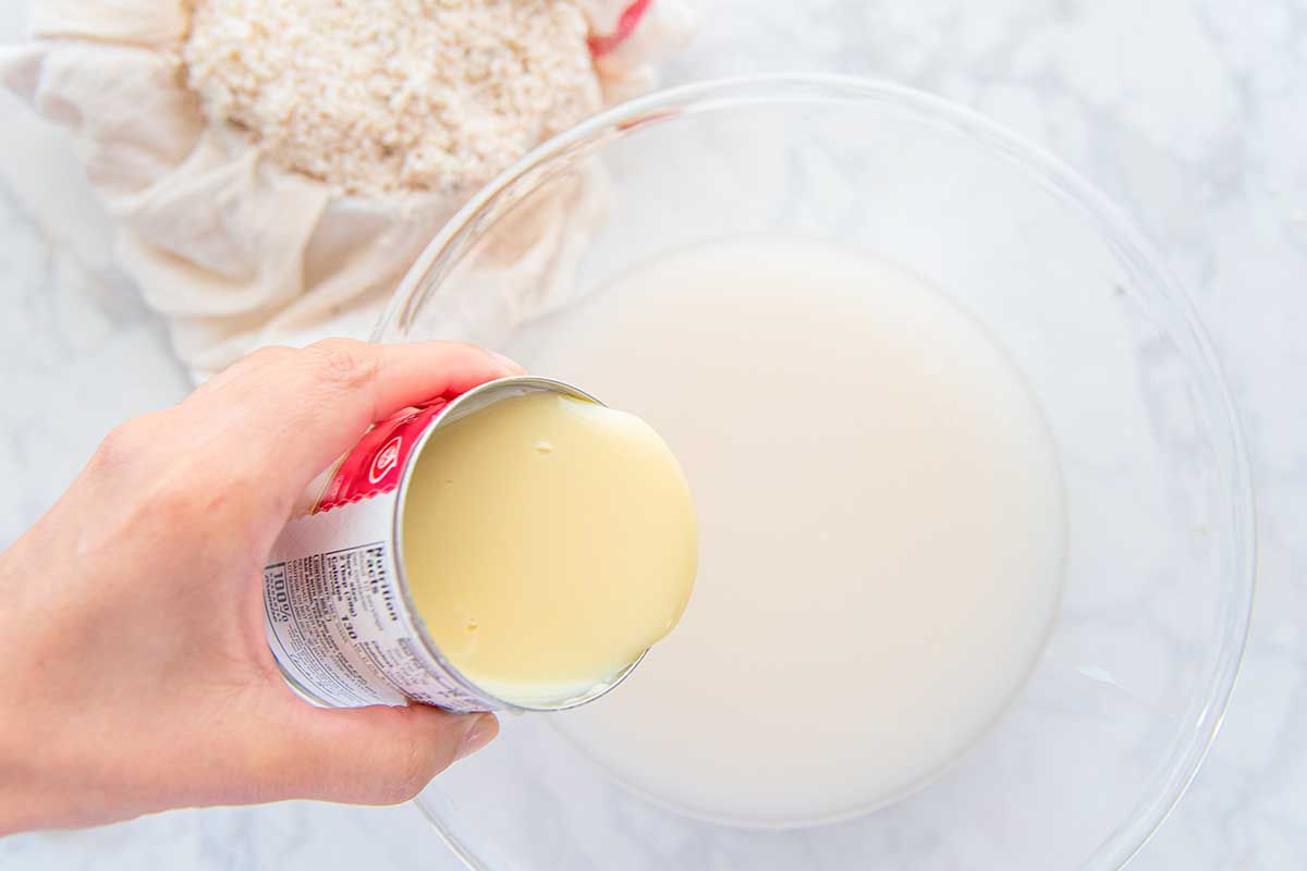 A creamy liquid is in a glass bowl and sweetened condensed milk is being added to it to make an easy homemade horchata. White rice is in a cheesecloth behind it.