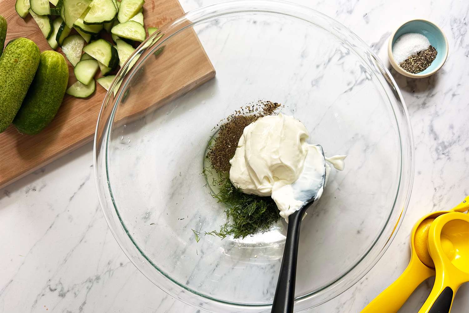 A bowl with yogurt herbs and spices surrounded by cucumbers and kitchen utensils