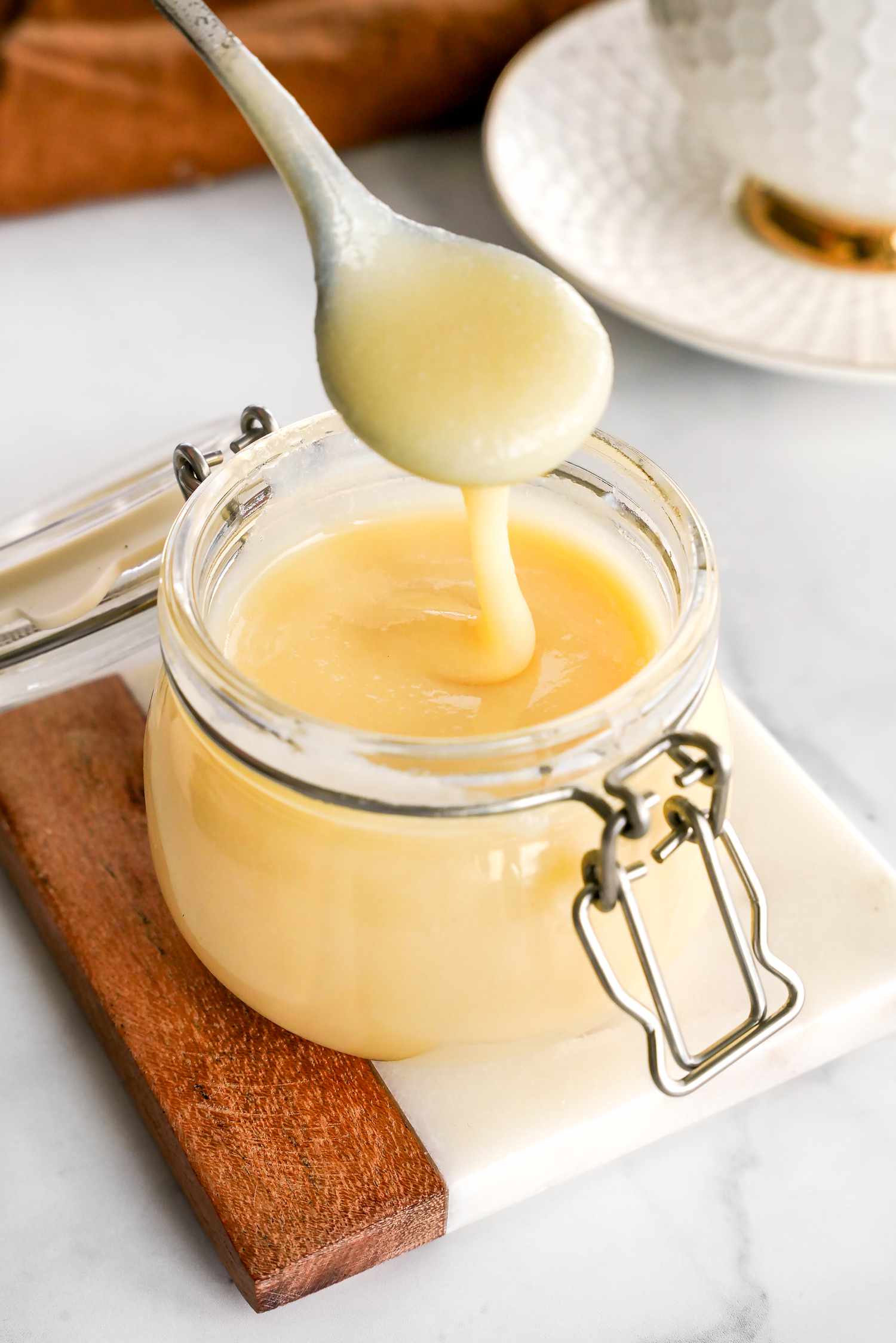 Spoonful of Homemade Condensed Milk Lifted from a Jar on a Coaster, and in the Background, a Kitchen Towel and a Teacup