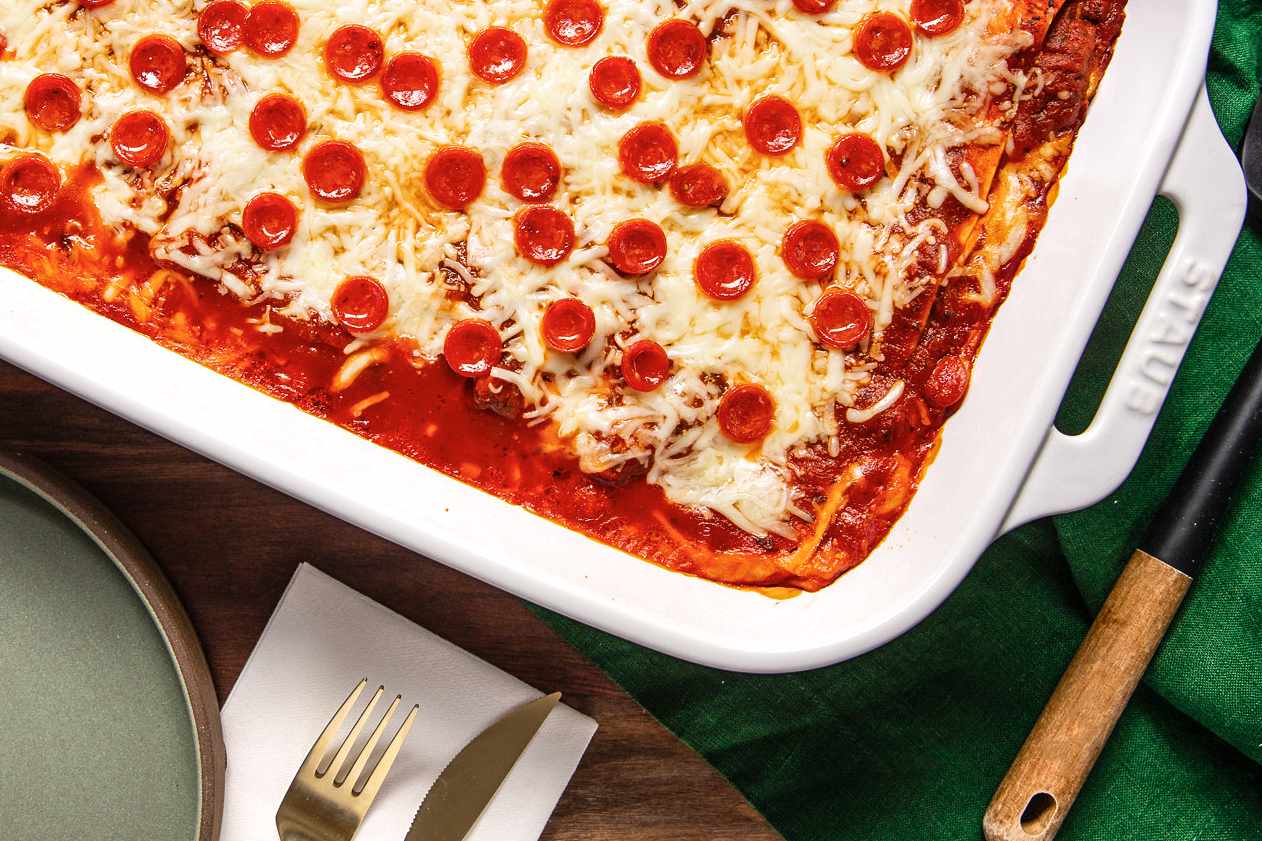 Pepperoni Pizza Lasagna in a Casserole Dish on a Green Kitchen Towel at a Table With a Plate, a Set of Utensils on a Napkin, and a Spatula on the Table