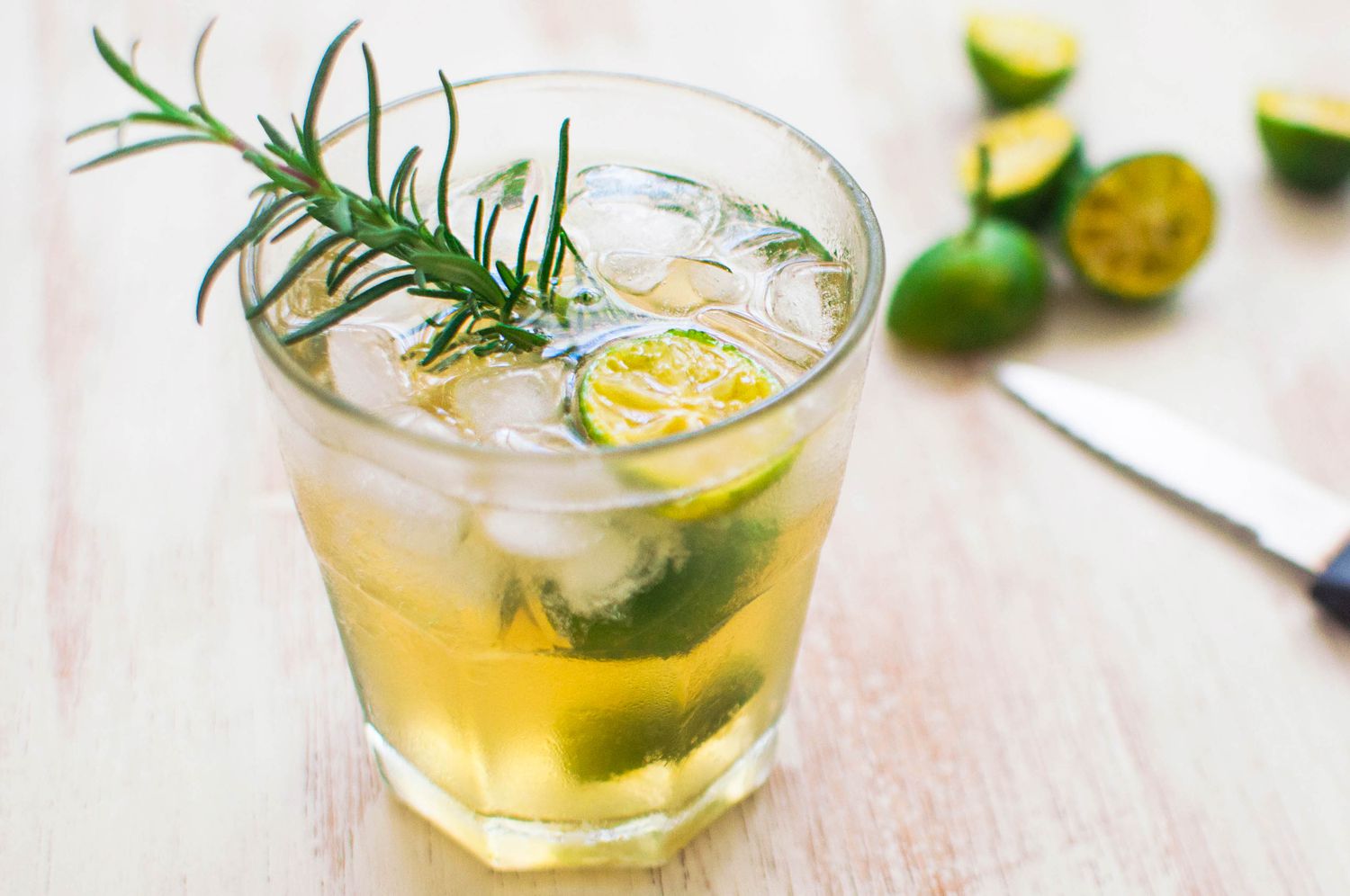A Calamansi Drink With Ice, a Rosemary Sprig, and a Halved Calamansi, and in the Background, Halved Calamansi on the Table
