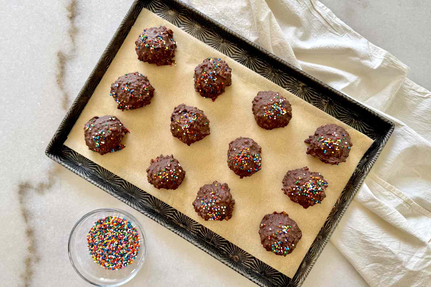 A tray of chocolate treats topped with sprinkles placed on parchment paper with a small bowl of sprinkles nearby