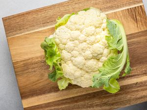 Whole cauliflower head on a wooden cutting board partially surrounded by green leaves