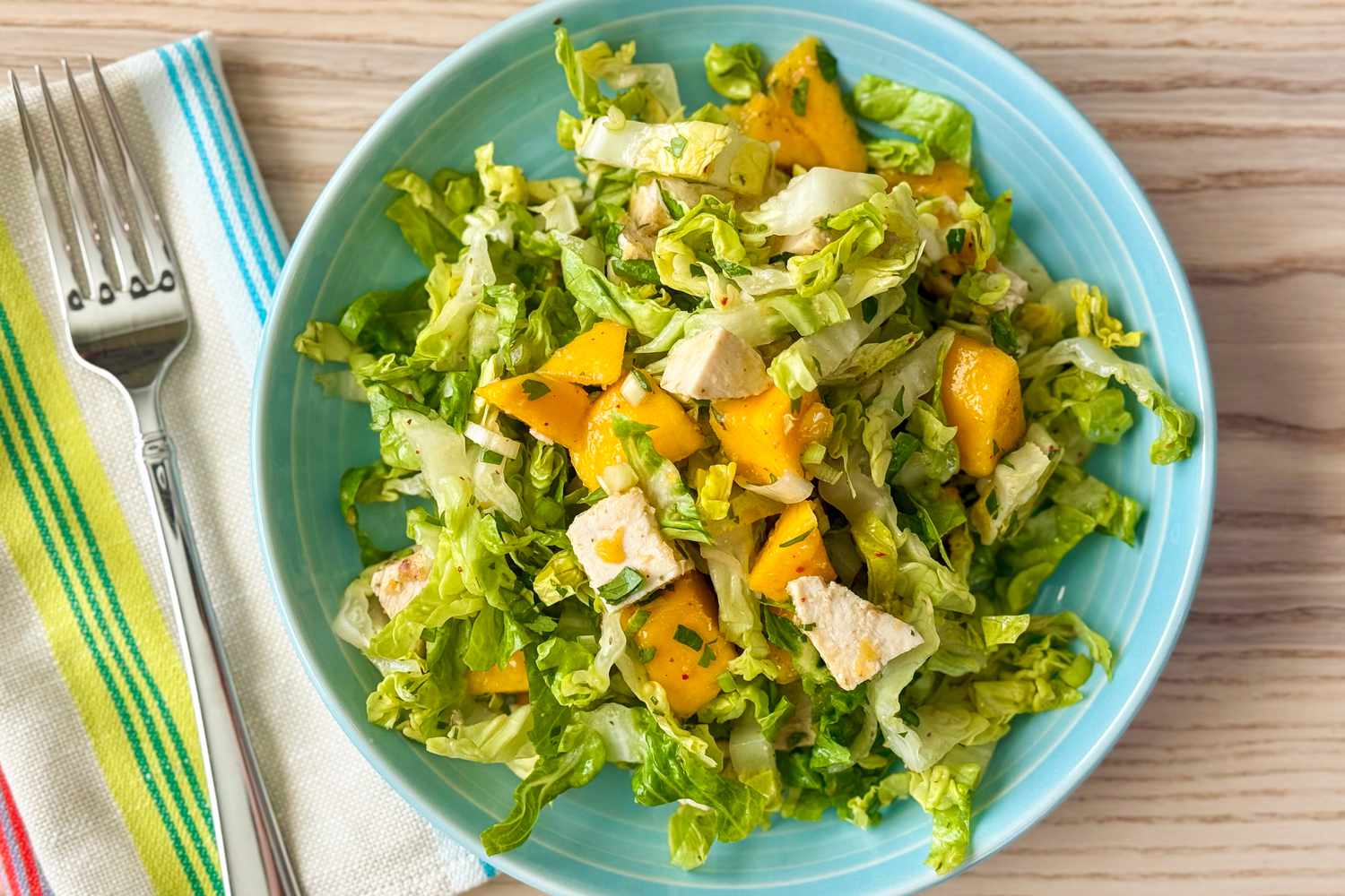 A plate of salad with lettuce, mango, and chicken cubes on a wooden surface with utensils beside it
