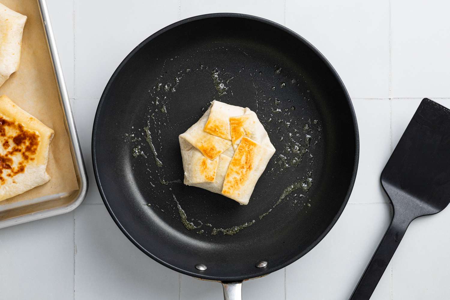 S'mores Crunchwrap Toasting on a Buttered Skillet, and Next to It, a Parchment Paper Lined Baking Sheet With More Crunchwraps and a Plastic Spatula 