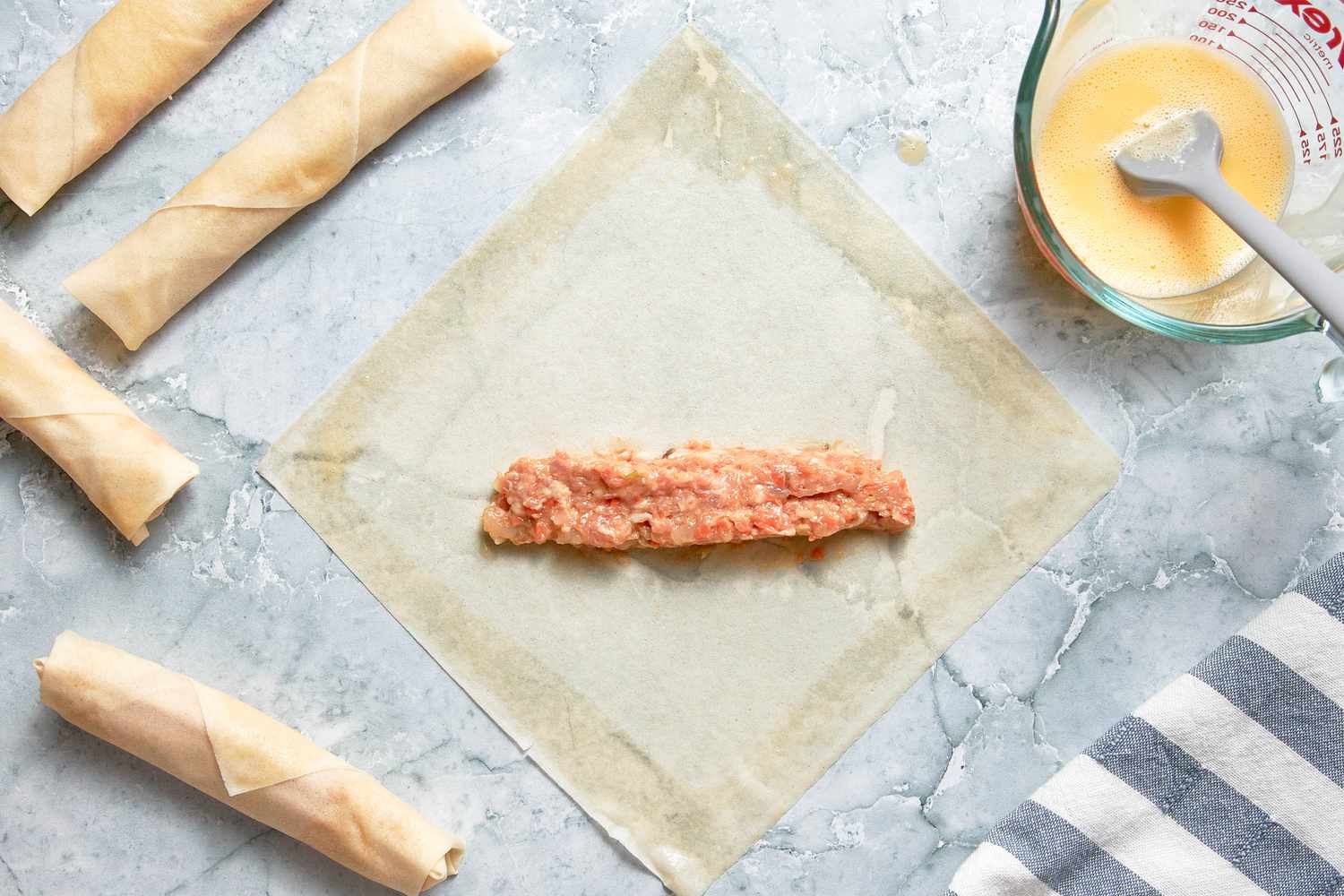 One Heaping Tablespoon of Filling in the Middle of an Egg Roll Wrapper With the Edges Brushed With Egg Wash, and in the Surroundings, and in the Surroundings, Rolled Lumpia, a Pyrex Measuring Cup With the Egg Wash, and a Kitchen Towel