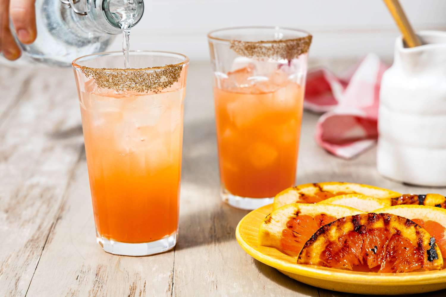 Clun Soda Poured Into a Salt Rimmed Glass of Grapefruit Paloma, and in the Surroundings, Another Glass of Paloma, a Cream Pitcher, and a Small Plate With Grilled Grapefruit Slices