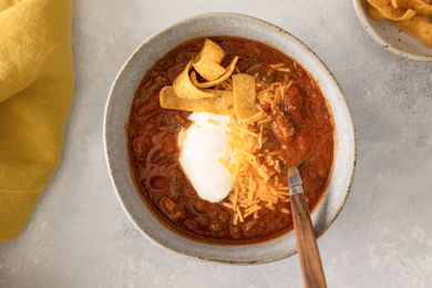 Bowl of Grandma Patty’s Chili with sour cream shredded cheese and Fritos alongside a bowl of Fritos and a yellow napkin