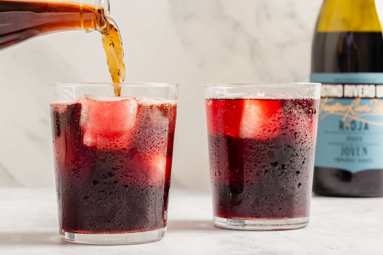 Two glasses filled with a red beverage, one being poured, with a bottle of wine nearby