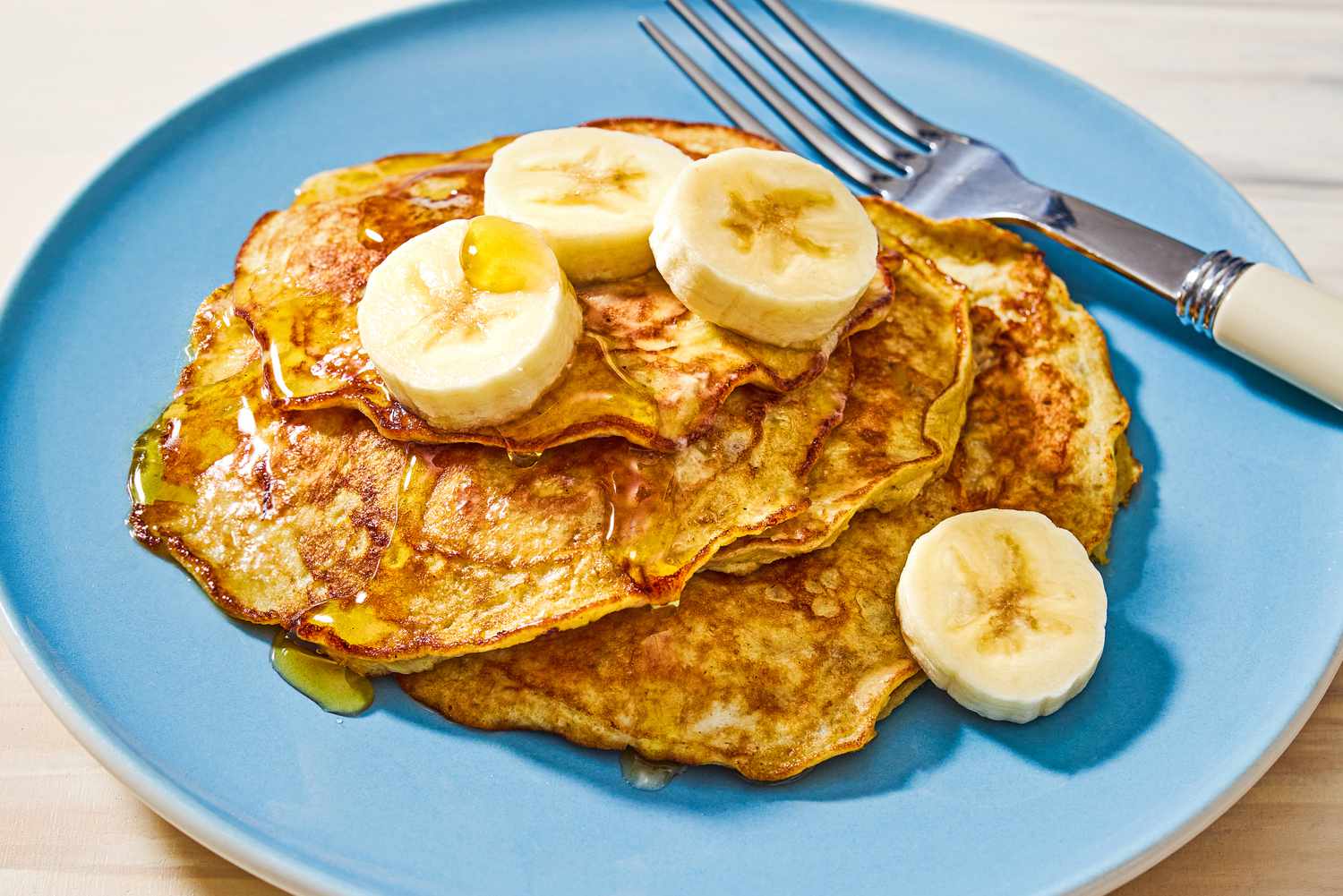Closeup view of a blue plate of 2-ingredient pancakes topped with bananas next to a fork, all on a wooden tabletop