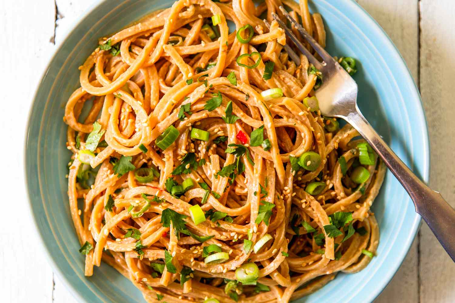 A plate of noodles with a peanut sauce, garnished with green onions and parsley, a fork placed on the side