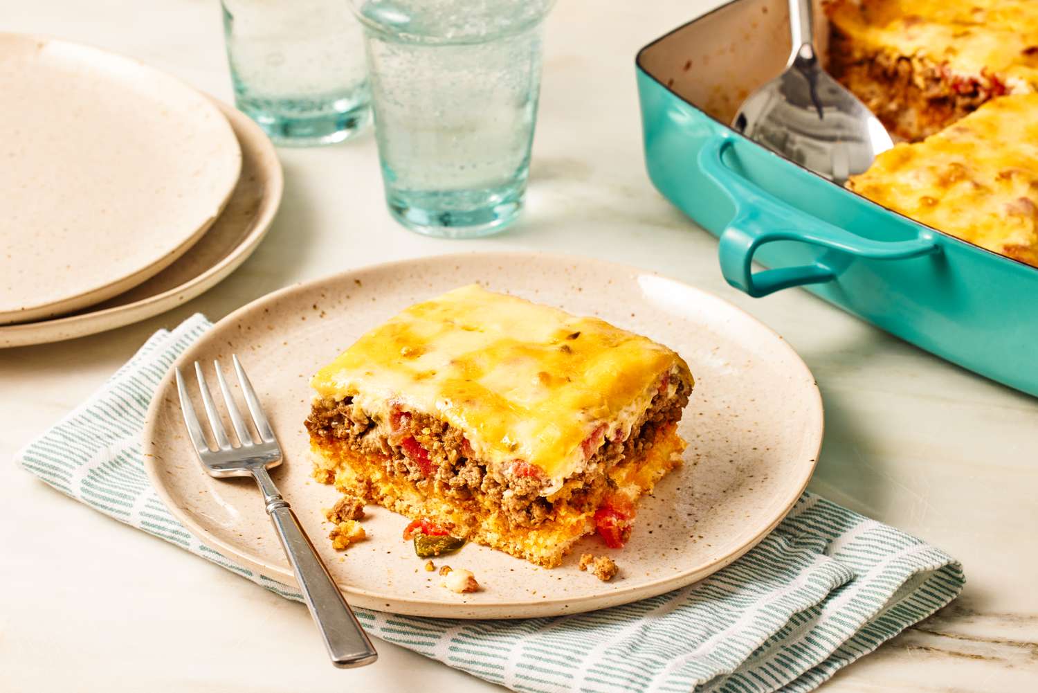 slice of cattle drive casserole on a plate with a fork at a table setting with a stack of plates, glasses of water, and more casserole in a casserole dish