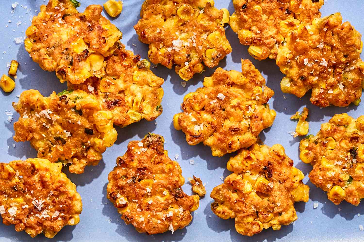 Corn fritters arranged on a flat surface and sprinkled with salt, consisting of fried mixtures with visible kernels