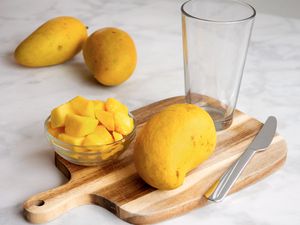 Sliced mango pieces in a bowl on a cutting board with a whole mango a glass and a knife nearby