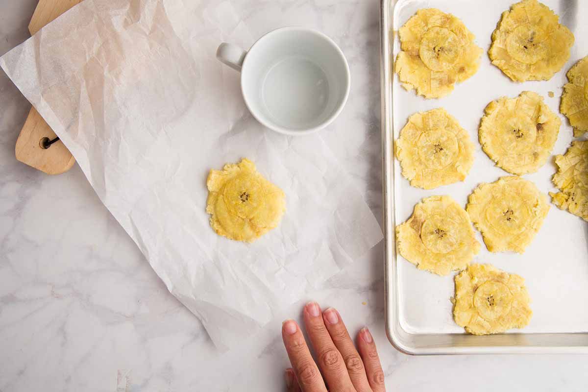 Smashing plantains (aka Tostones) between two pieces of parchment with a coffee mug and setting them on a parchment lined baking sheet.