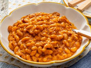 A bowl of baked beans with sauce and a spoon on a patterned tablecloth