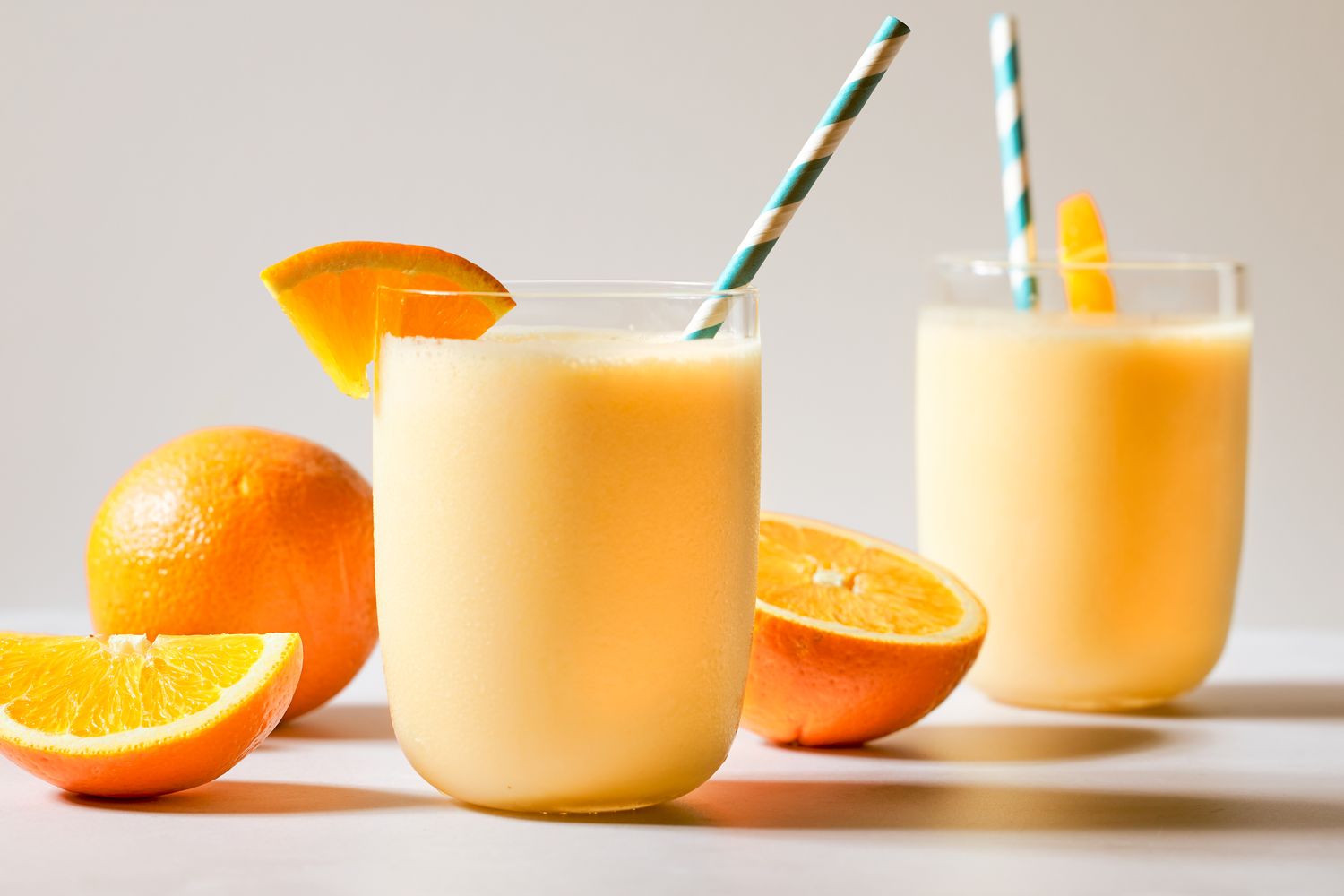 Two glasses of orange-flavored beverages with straws, garnished with orange slices, surrounded by whole and cut oranges